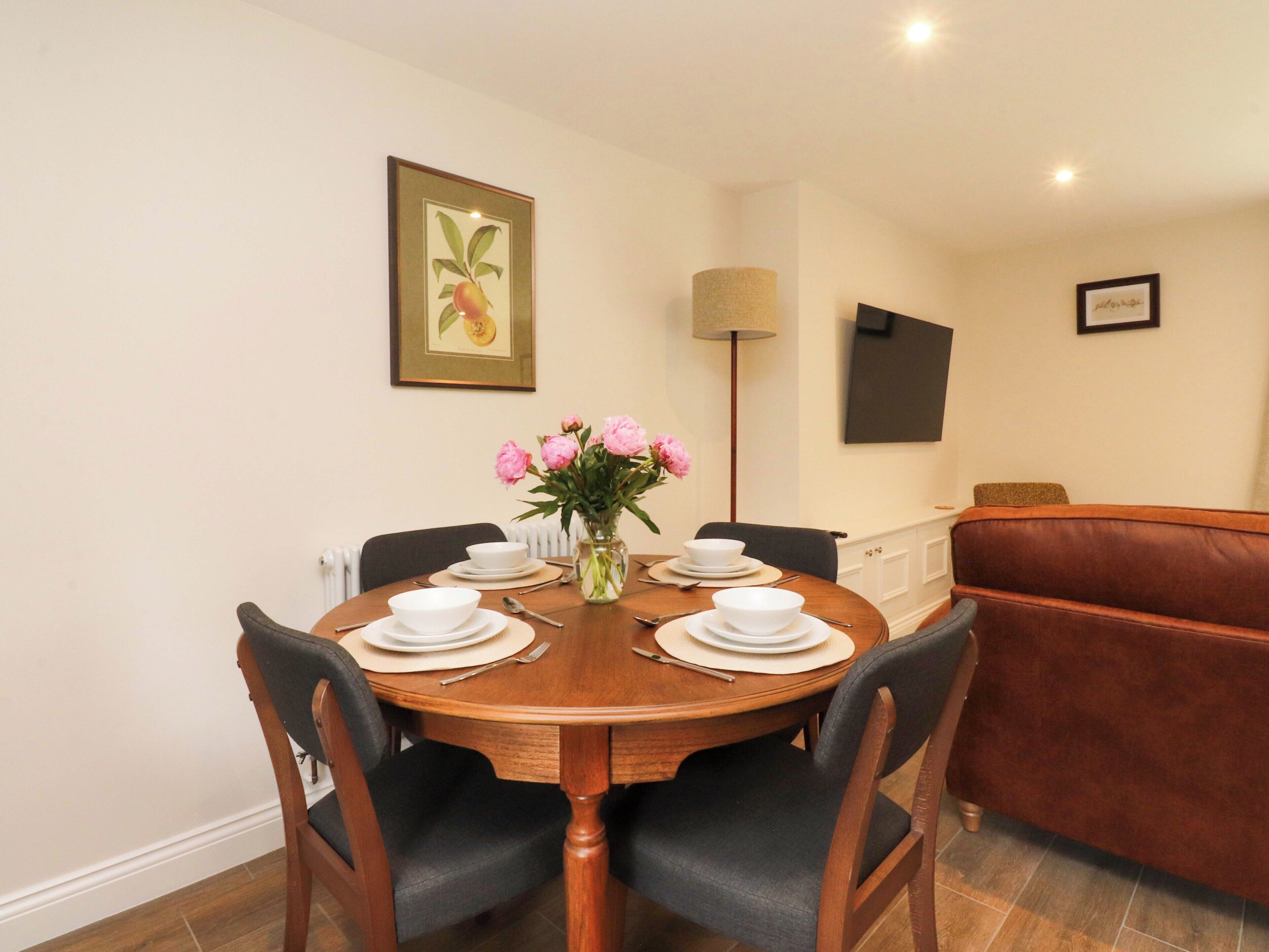 A dining area with a round wooden table set for four, surrounded by chairs. A vase with pink flowers is placed on the table.