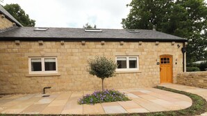 A stone building with a wooden door, two windows, and a small tree with purple flowers in the center. - Heron View (Preston)
