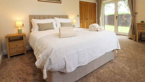 A bedroom with a large bed, two bedside tables with lamps, a wooden door, and a view through a glass door. - Heron View (Preston)