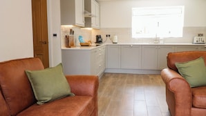 A modern kitchen with white cabinets, a countertop with kitchen appliances, and a window providing natural light. There are two brown leather sofas with green cushions. - Heron View (Preston)