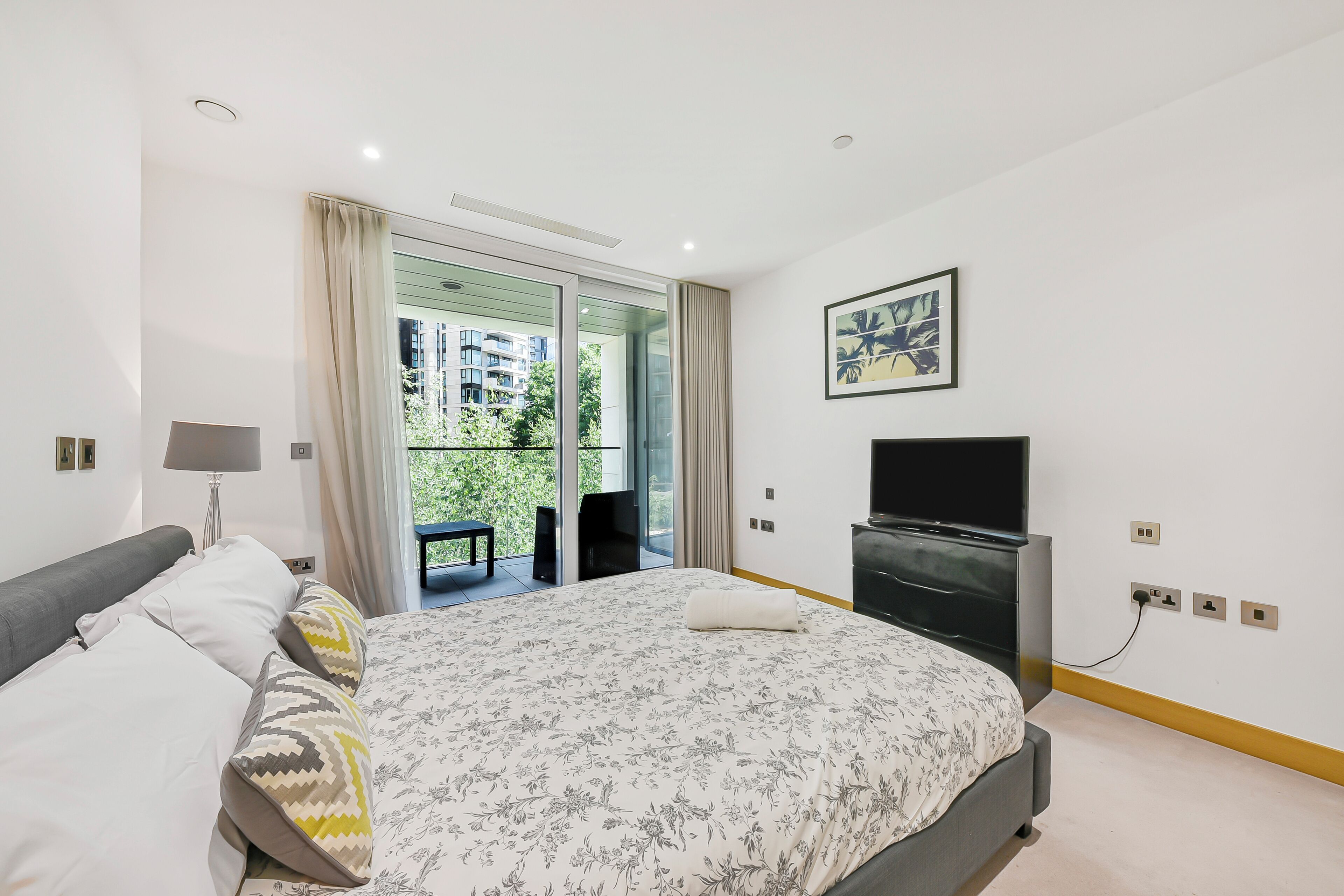 A modern bedroom with a large bed, a flat-screen TV, and a balcony with a view of buildings and greenery.