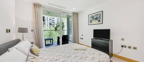 A modern bedroom with a large bed, a flat-screen TV, and a balcony with a view of buildings and greenery.