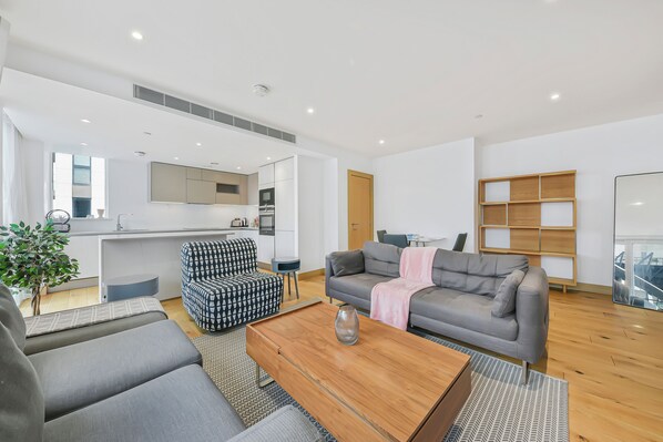 A modern living room with a grey sofa, a patterned armchair, and a wooden coffee table. A kitchen area is visible in the background with a built-in oven and microwave. - Paddington Ex LUX Flats (London)