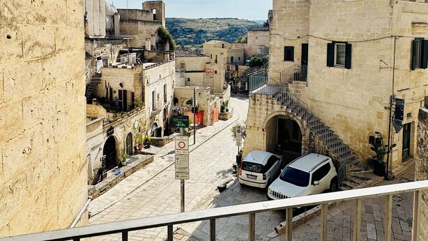 A view from a balcony overlooking a narrow street with stone buildings, a parked car, and a clear sky. - Il Sorriso dei Sassi Rooms (Matera)