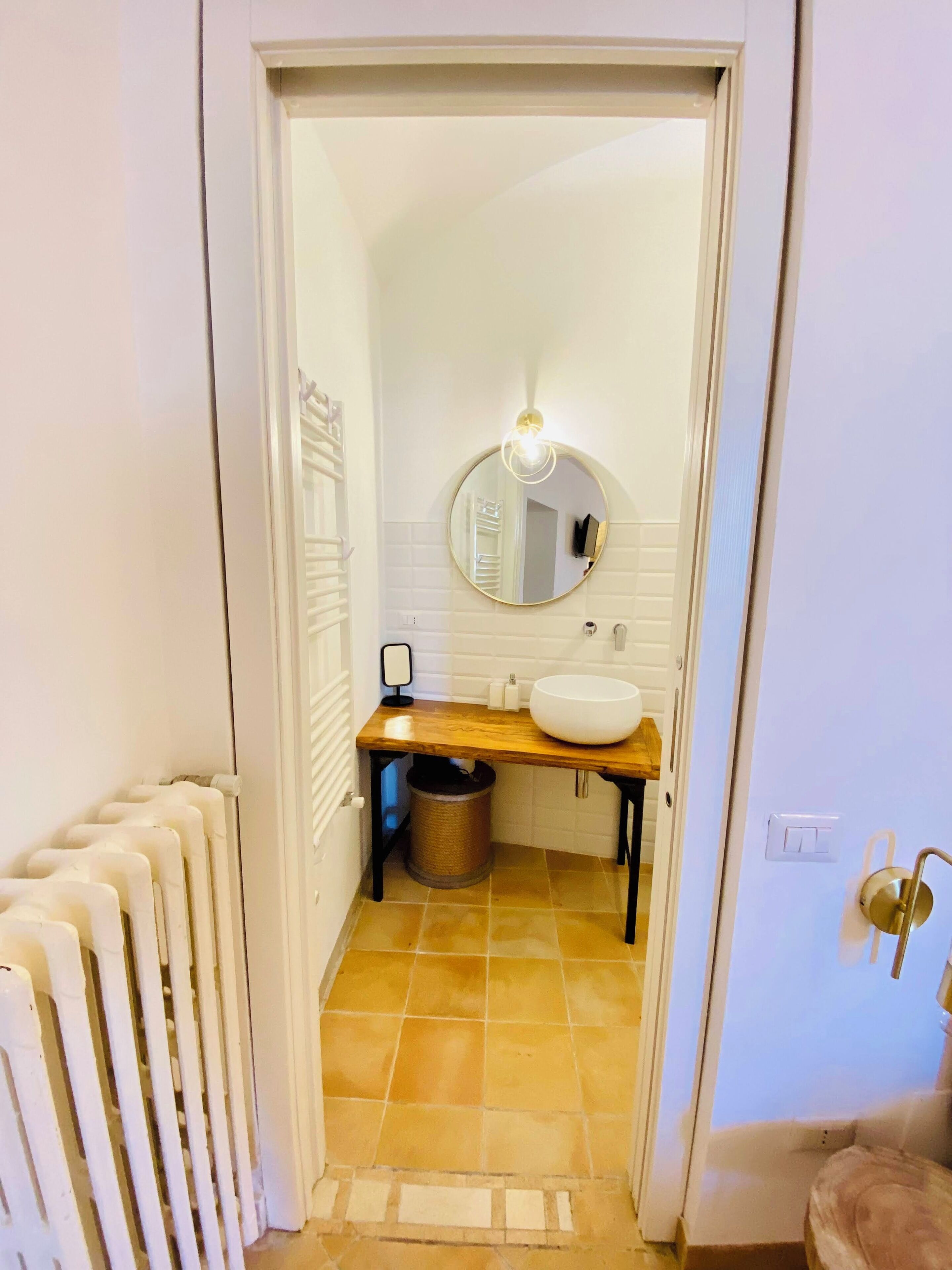 Un couloir étroit mène à une salle de bain équipée d’un lavabo blanc, d’un miroir rond et d’un luminaire placé au-dessus.