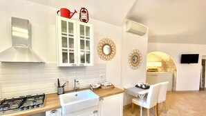 A modern kitchen with white cabinets, a gas stove, and a sink. There is a dining area with a table and chairs, and a TV mounted on the wall. - Il Sorriso dei Sassi Rooms (Matera)