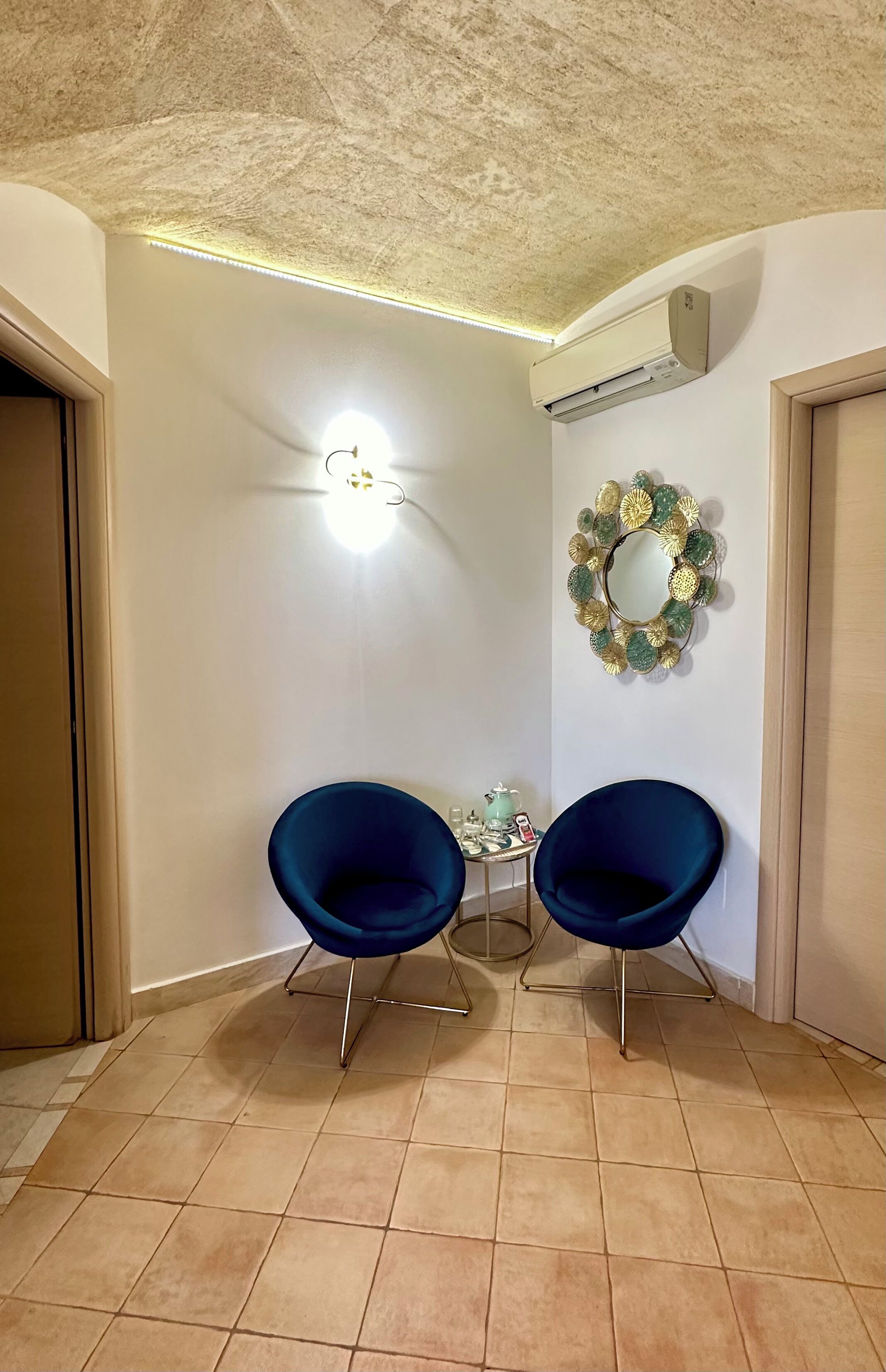 Quarto com duas cadeiras azul-escuras, uma mesinha com objetos decorativos, um espelho fixado na parede e um aparelho de ar condicionado.