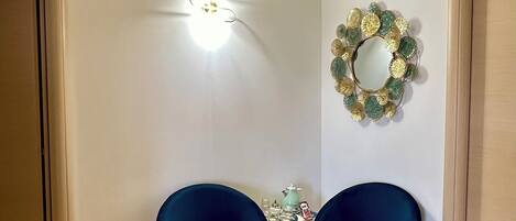 Quarto com duas cadeiras azul-escuras, uma mesinha com objetos decorativos, um espelho fixado na parede e um aparelho de ar condicionado.