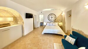 A spacious bedroom with a large bed, a television, a clock, and a stone wall. - Il Sorriso dei Sassi Rooms (Matera)