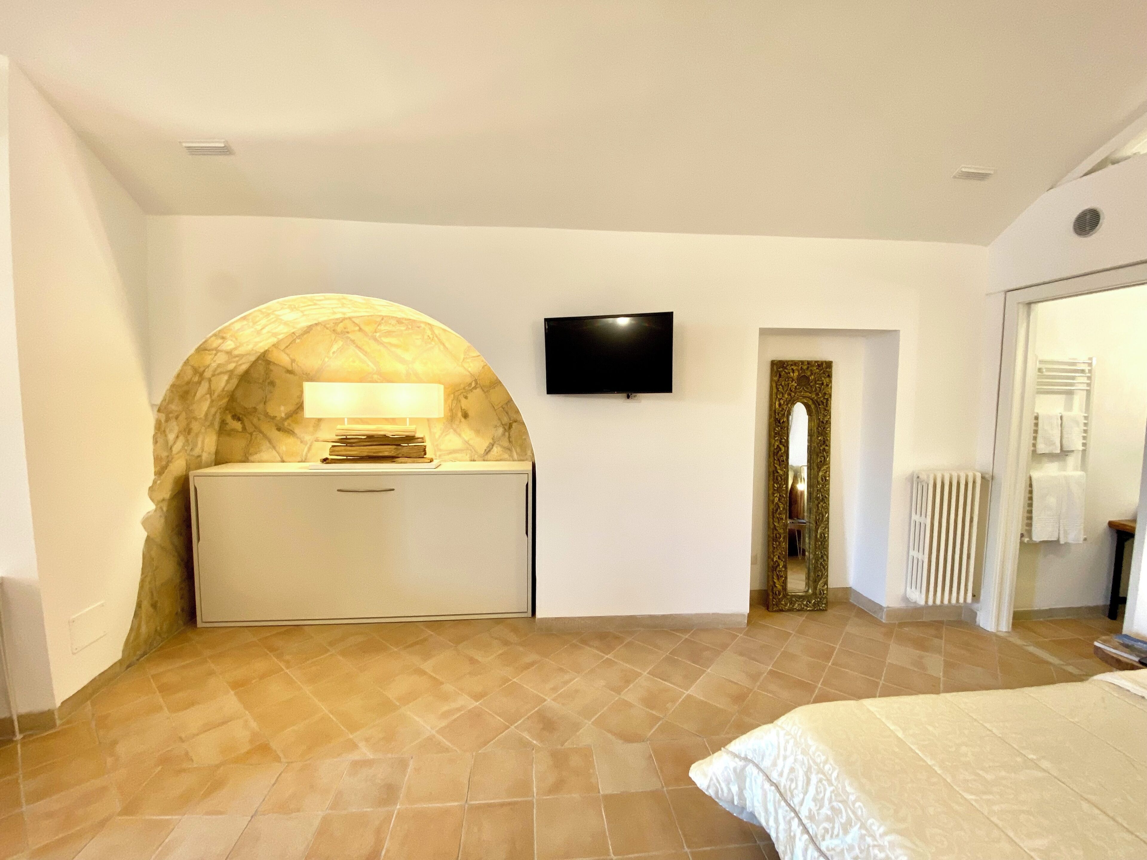 A bedroom with a bed, a TV, a desk with a lamp, and a bathroom with a radiator.
