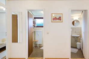 Un couloir comprenant un lit, un radiateur, un tabouret, un tableau encadré, ainsi qu’une salle de bain avec un lavabo et des toilettes.