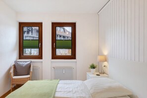 Un dormitorio con una cama, una silla, una mesita de noche con lámpara y dos ventanas con persianas.