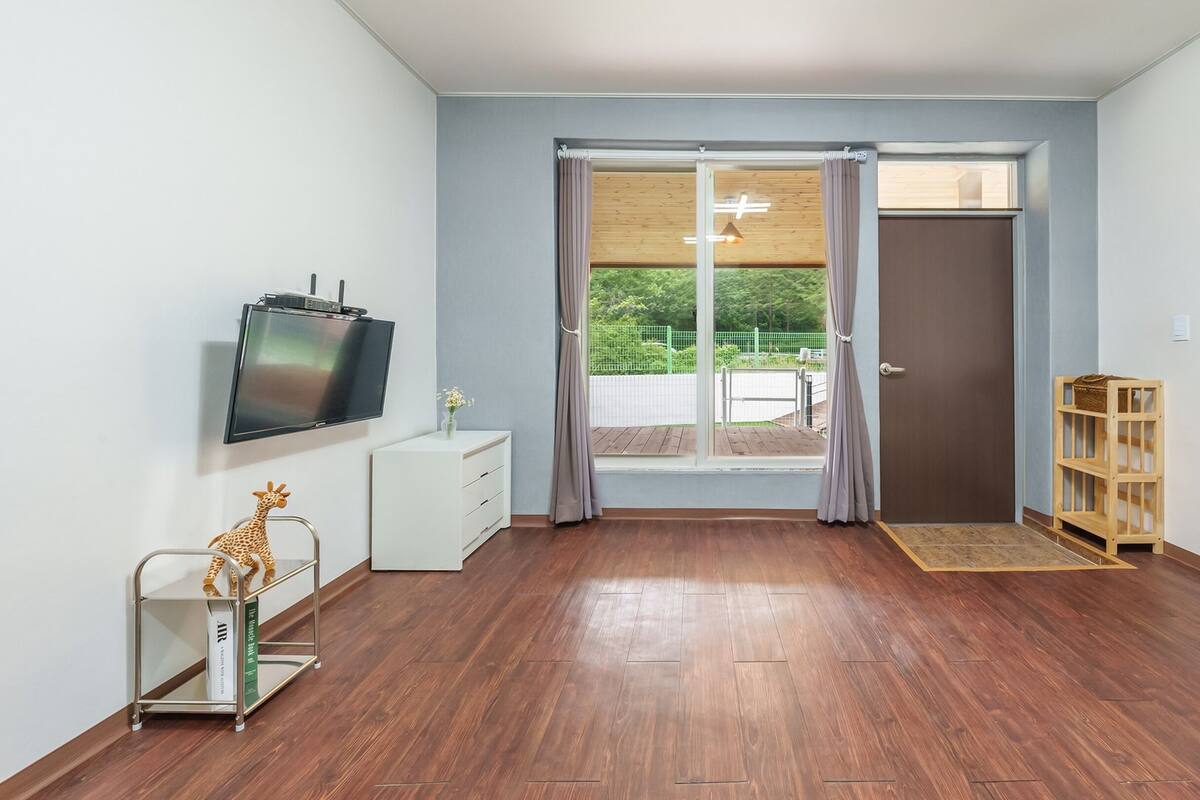 Une chambre moderne avec un plancher en bois, un téléviseur à écran plat, une commode blanche, un fauteuil rose, une bibliothèque en bois et une porte-fenêtre coulissante donnant sur un espace extérieur.
