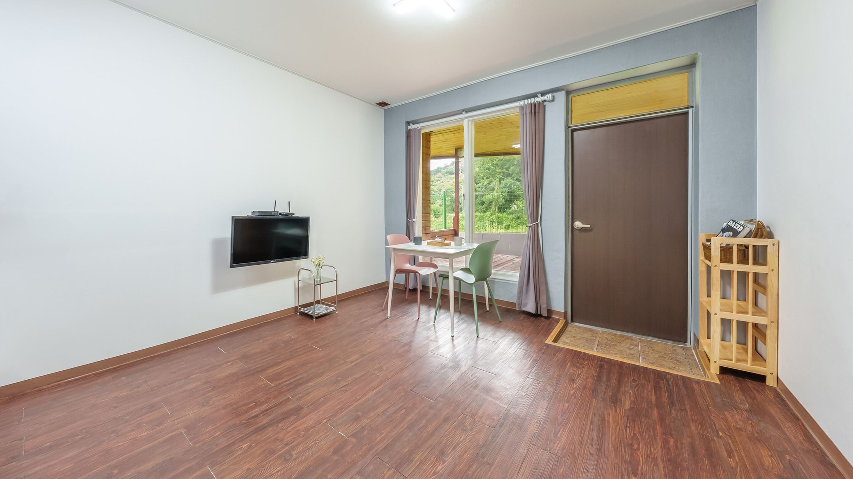 Une pièce avec un plancher en bois, un téléviseur fixé au mur, une table avec des chaises, une fenêtre avec des rideaux et une porte.