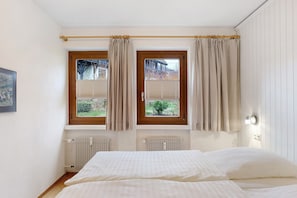 Quarto com duas janelas, uma cama, um radiador e um quadro na parede.