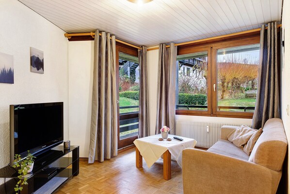 A modern living room with a flat-screen TV, a sofa, a small table with a vase, and large windows with curtains. - Ferienwohnung 2 mit Balkon – barrierefrei, 2 Schlafzimmer, Küche, Bernau im Schwarzwald (Bernau im Schwarzwald)