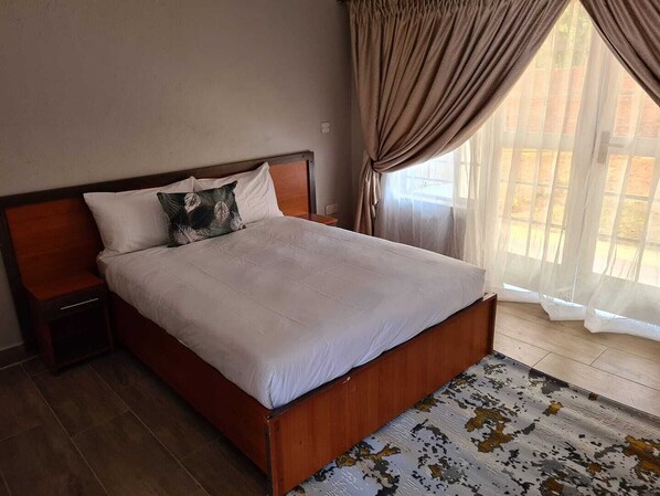 A neatly made bed with white linens and a wooden headboard, a nightstand with a lamp, and a patterned rug on the floor. - Blissful lily lodge (Salima)