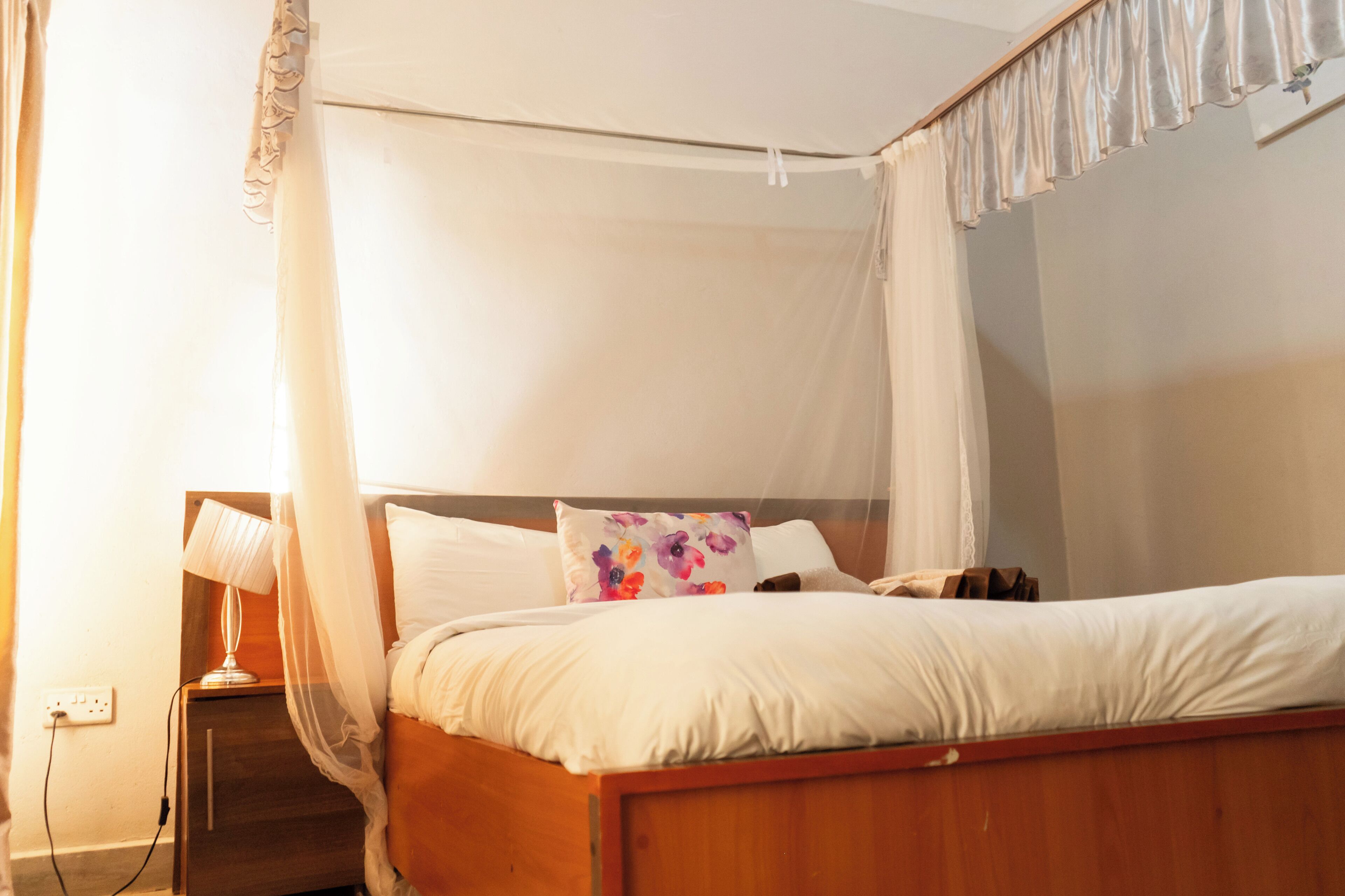 Un dormitorio con cama de madera, ropa de cama blanca, una lámpara de noche y un enchufe.