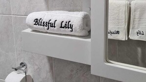 A bathroom with a toilet, a towel rack with a roll of toilet paper, and a shelf with a folded towel labeled "Blissful Lily". - Blissful lily lodge (Salima)