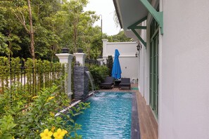 A swimming pool with a waterfall feature, surrounded by lush greenery and a white building with green window frames. - Fortune Home Luxury Villa Kem Beach (Phu Quoc)