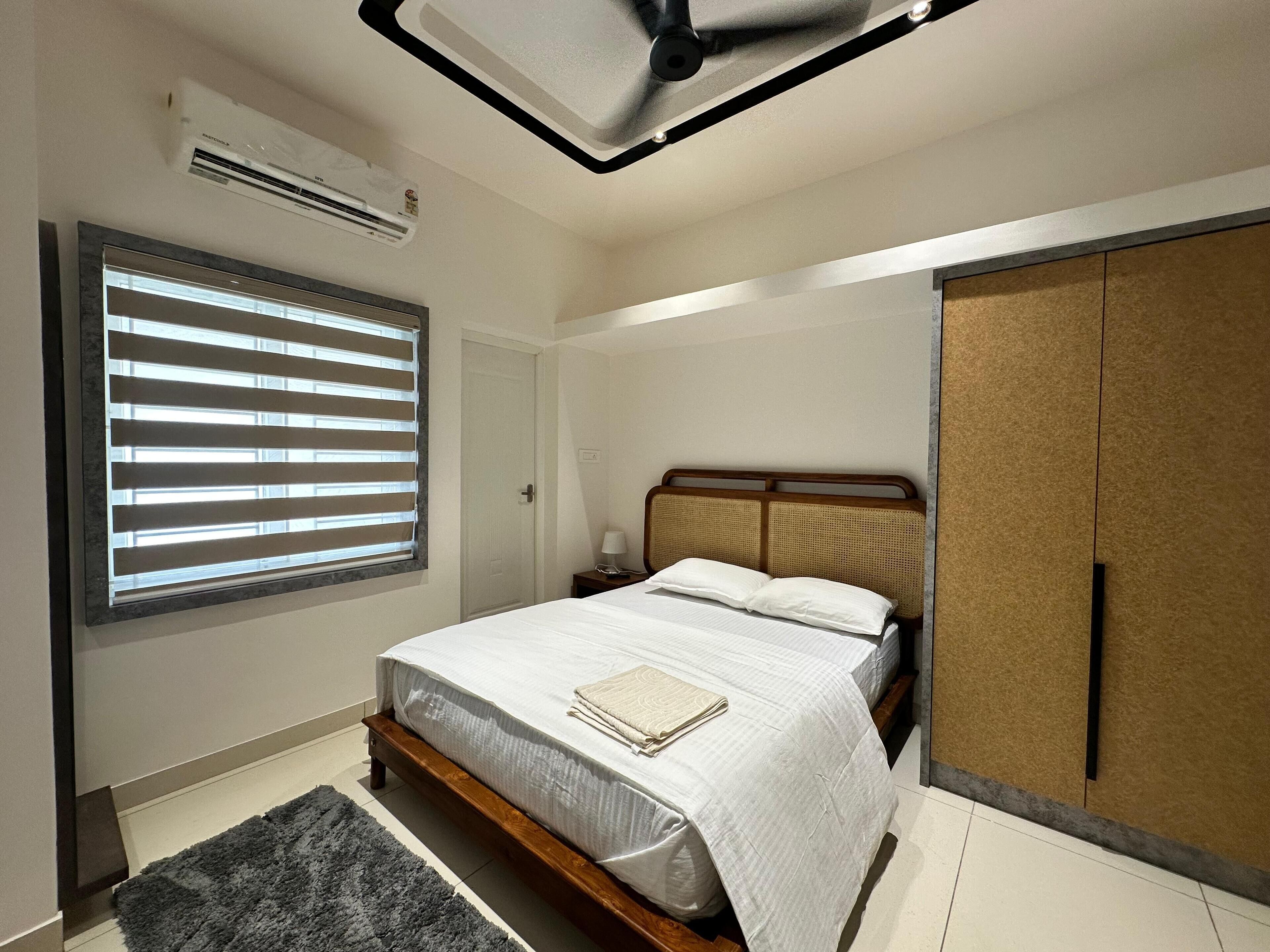 Un dormitorio con cama, ventana con persianas, aire acondicionado, mesita de noche con lámpara y armario.