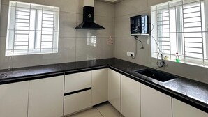 A kitchen with white cabinets, a black countertop, a black range hood, and two windows with white blinds. - Thulasi Avenue (Pollachi)