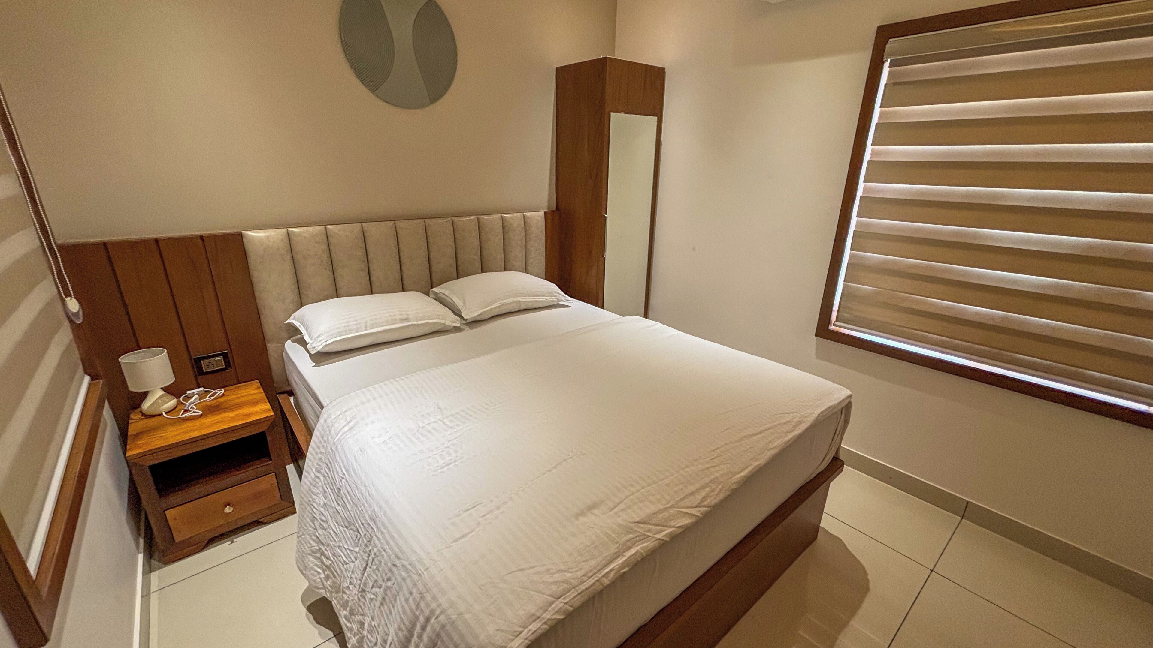 Une chambre à coucher comprenant un lit, une table de chevet, un miroir et une fenêtre avec des stores.