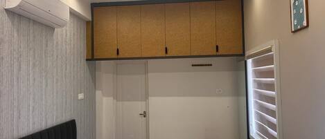 A bedroom with a bed, a ceiling fan, an air conditioning unit, a wardrobe, a window with blinds, and a small table.