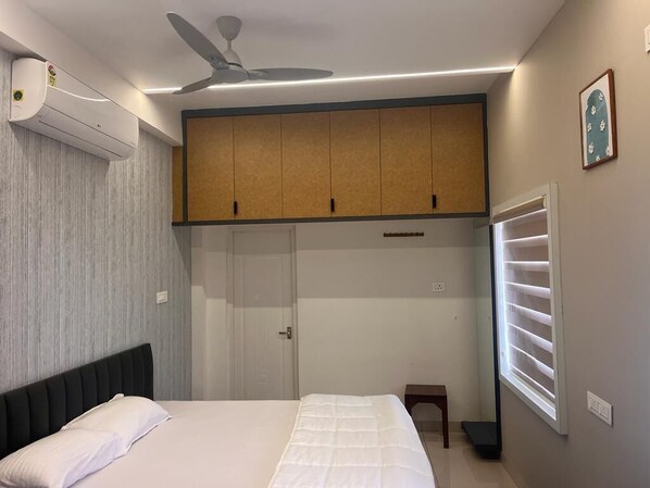 A bedroom with a bed, a ceiling fan, an air conditioning unit, a wardrobe, a window with blinds, and a small table. - Thulasi Avenue  (Pollachi)