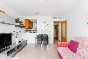 A modern living room with a flat-screen TV, a dining table with chairs, a sofa with a patterned throw, and a kitchen in the background.