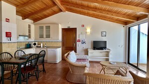 Une cuisine compacte avec des meubles en bois, un coin repas avec des chaises, un canapé en osier et un téléviseur posé sur un meuble.