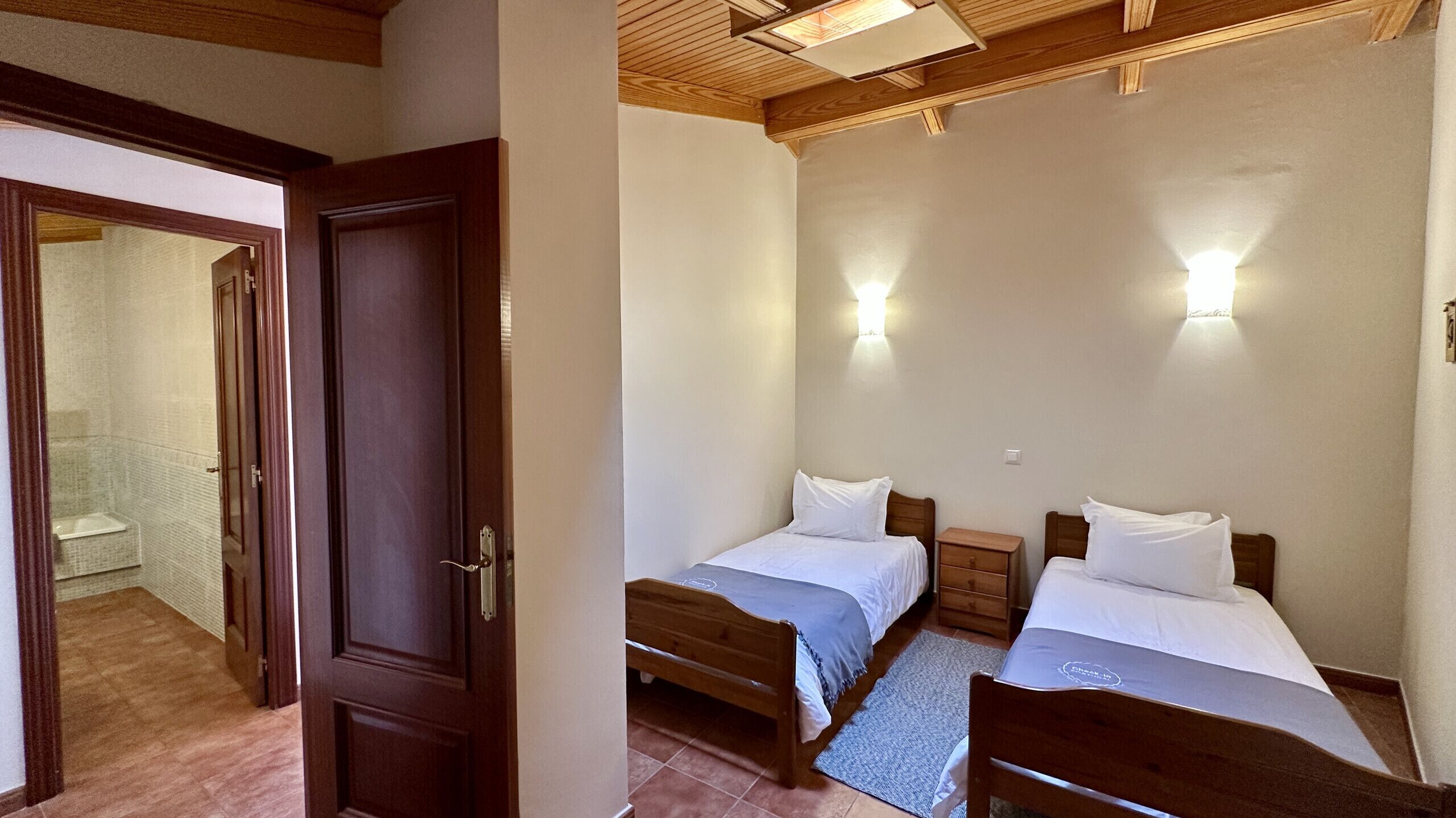 Un dormitorio con dos camas individuales, una puerta de madera, un baño con ducha, y un techo de vigas de madera e iluminación empotrada.