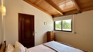 Une chambre avec un plafond en bois, une fenêtre avec des stores, une armoire en bois et un lit recouvert d’une couverture blanche.