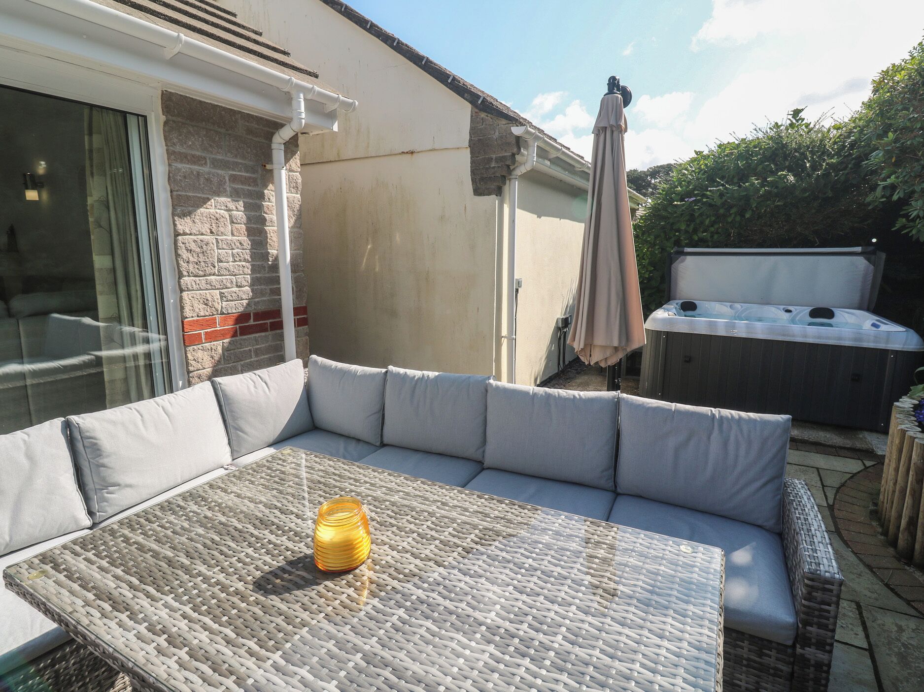 A patio with a wicker table, a cushioned sofa, a hot tub, and a glass door leading to a room.