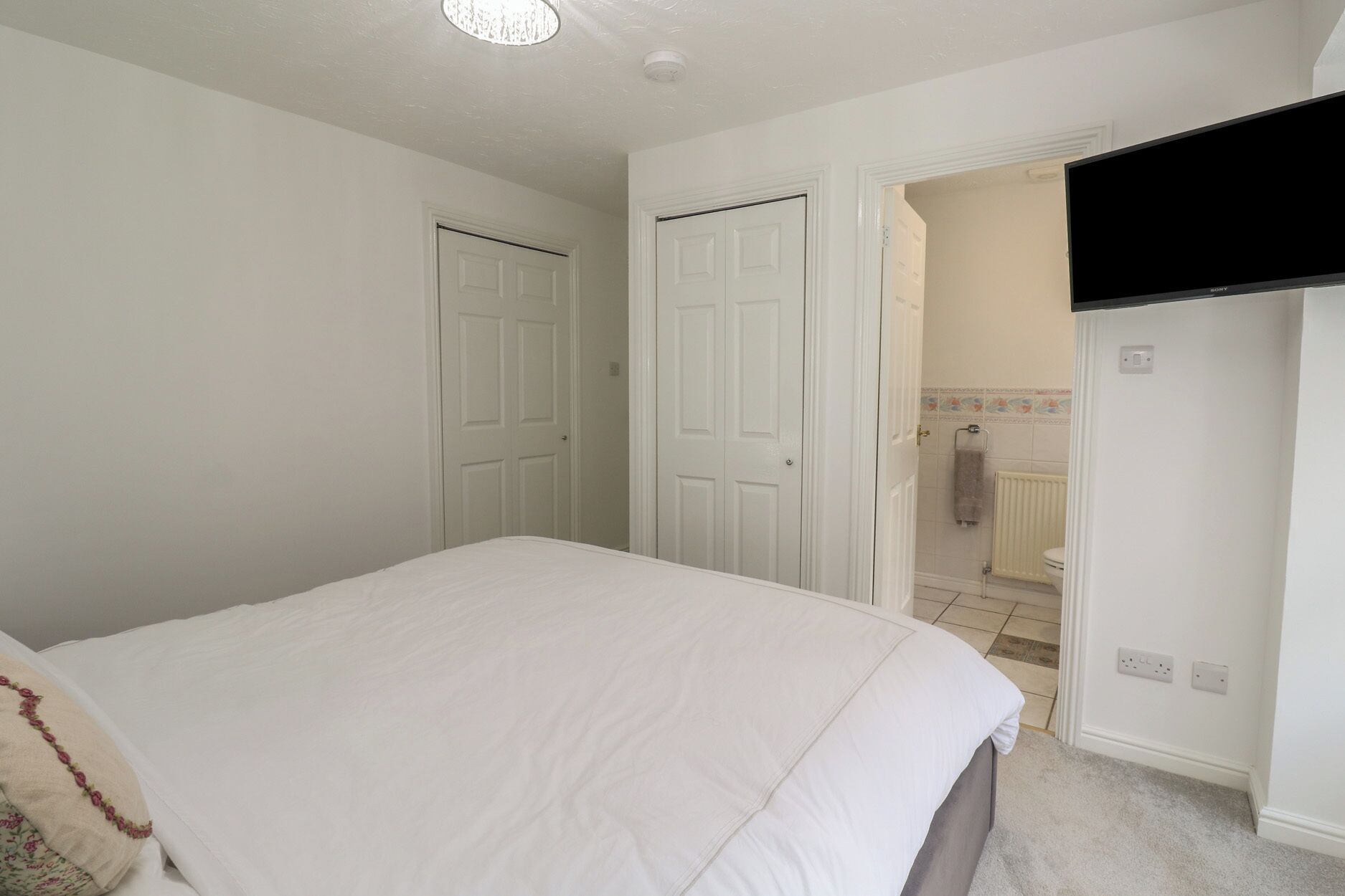 A bedroom with a bed, two white doors, a bathroom with a toilet and shower, and a flat-screen TV mounted on the wall.