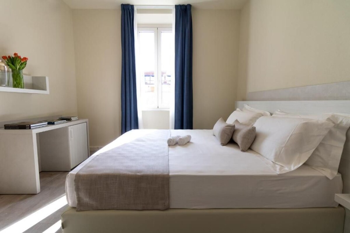 Un letto rifatto con cuscini e coperta, un comodino con libri, una piccola mensola con fiori e una finestra con tende.