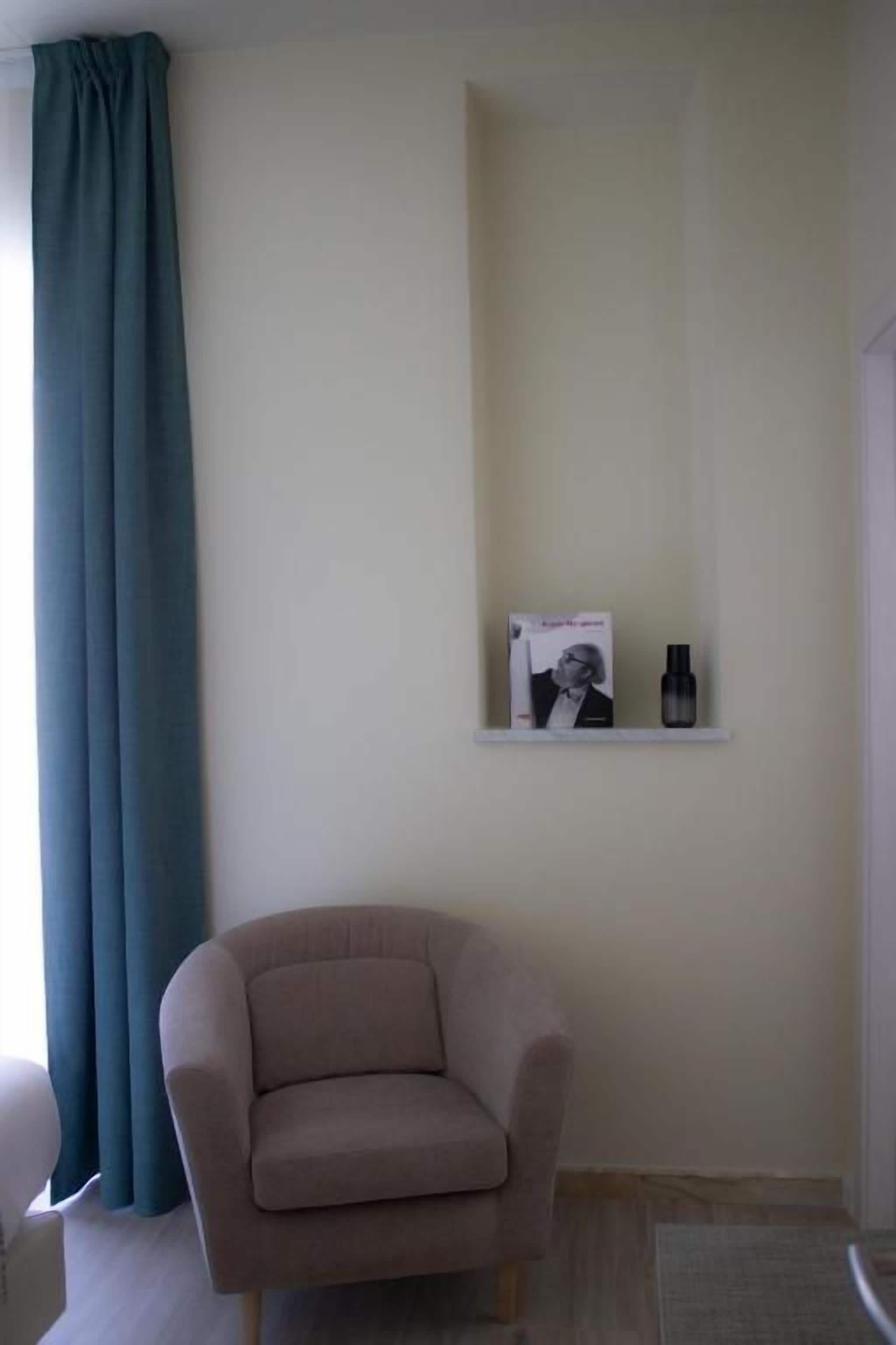 Ein Zimmer mit einem beigefarbenen Sessel, einem türkisfarbenen Vorhang, einem weißen Regal mit einem gerahmten Bild und zwei Flaschen sowie einem Holzboden.