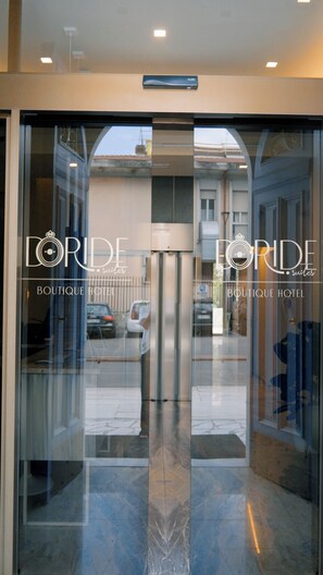 Fachada de un hotel con puertas de vidrio, un letrero que dice 'DORIDE Suites Boutique Hotel' y un reflejo del exterior.