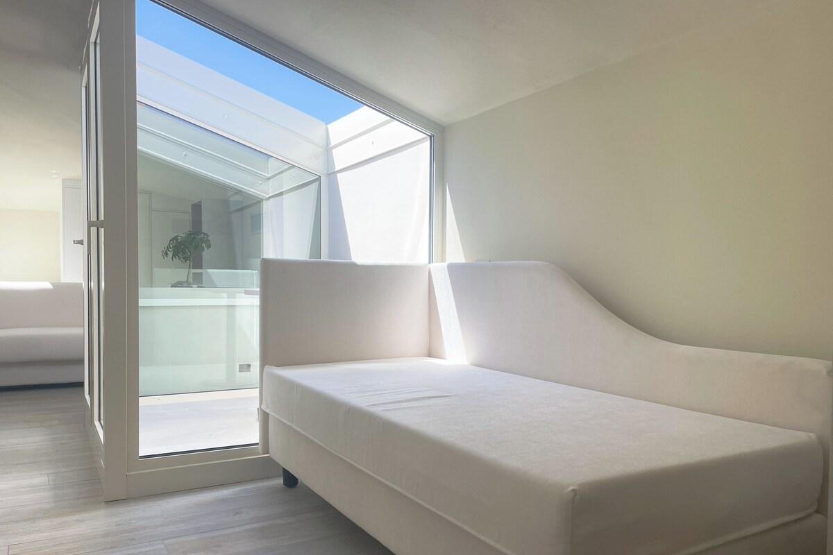 Una habitación moderna con un sofá blanco, un ventanal de vidrio a un balcón y suelo de madera.