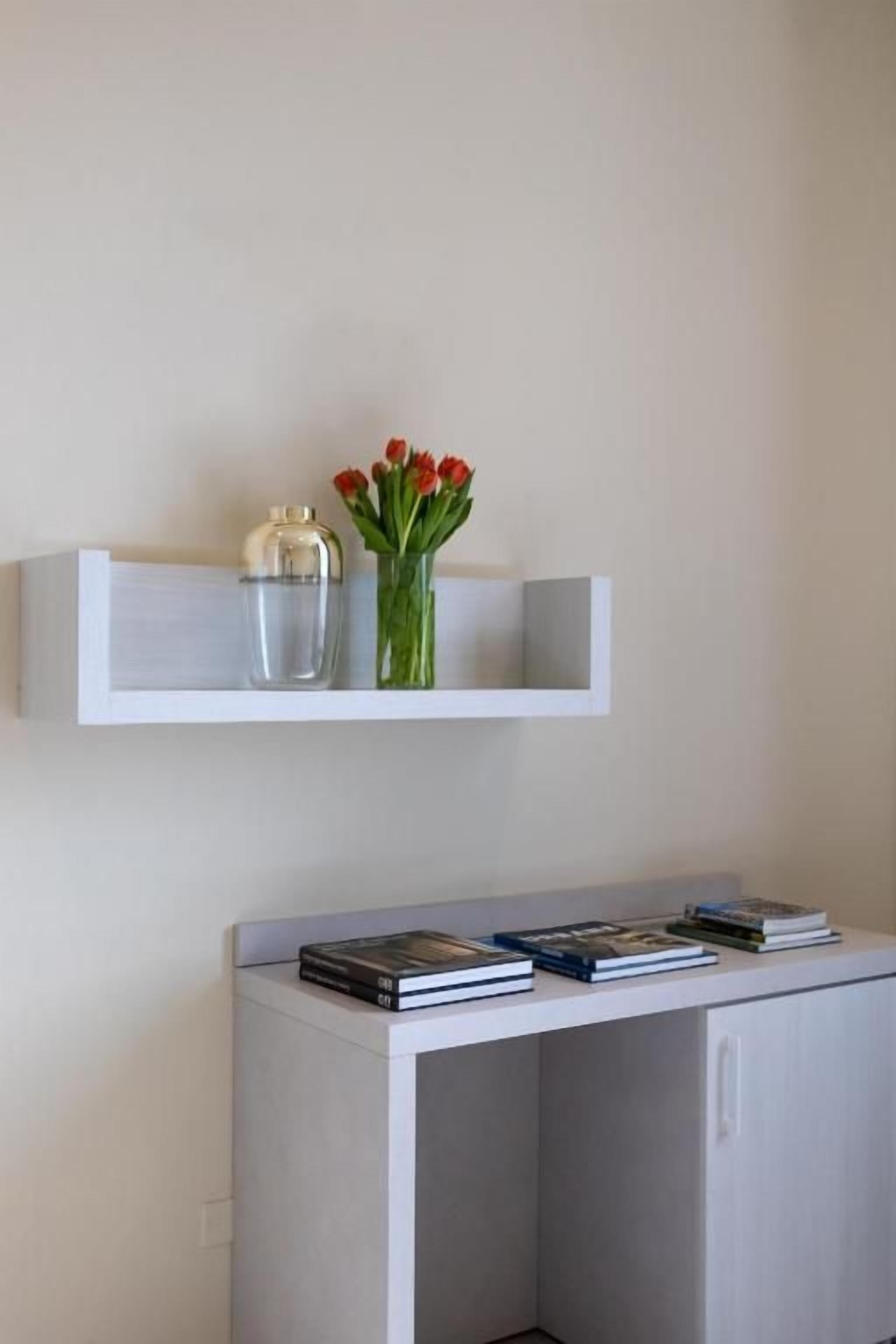 Una estantería blanca con un jarrón de flores rojas y un recipiente de vidrio transparente. Abajo, un armario blanco con libros y una mesita pequeña son visibles.