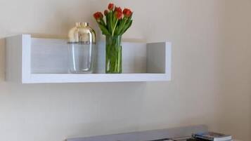 Una estantería blanca con un jarrón de flores rojas y un recipiente de vidrio transparente. Abajo, un armario blanco con libros y una mesita pequeña son visibles.