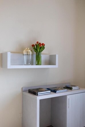 Una estantería blanca con un jarrón de flores rojas y un recipiente de vidrio transparente. Abajo, un armario blanco con libros y una mesita pequeña son visibles.