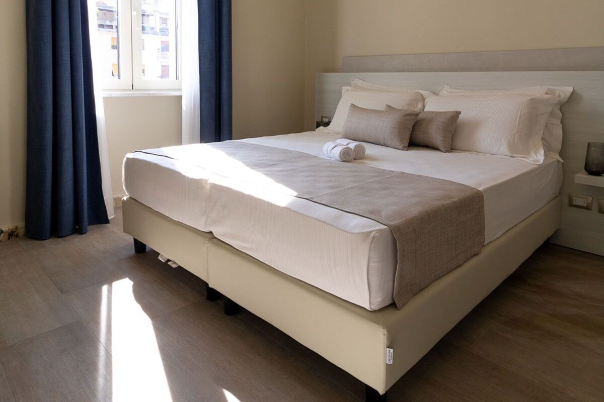 Una cama bien hecha con cabecera, una ventana con cortinas y una mesita de noche con lámpara.