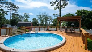 Une piscine circulaire entourée d’une terrasse en bois, un espace de détente couvert et un espace clos avec un barbecue.