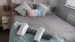 Un lit bien fait, avec une couette grise, des oreillers de différentes couleurs et une couverture turquoise, ainsi que des serviettes posées sur une table de chevet.