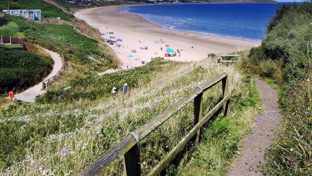 Un sentier côtier qui mène à une plage, bordée d’une clôture en bois, avec des zones herbeuses et une vue dégagée sur la mer.