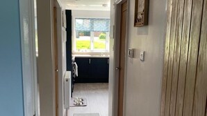 Un couloir étroit avec une porte en bois sur la droite, une fenêtre donnant sur un jardin et un bol pour chien posé sur le sol.