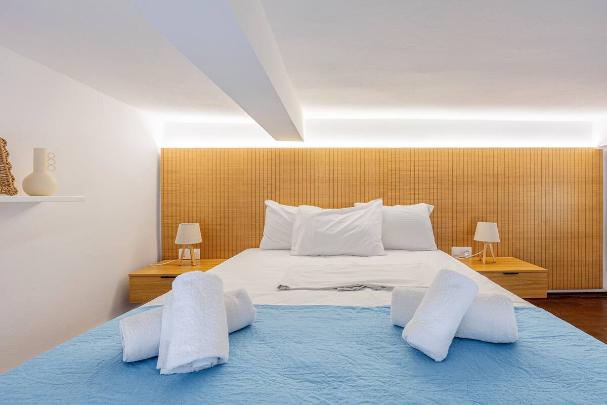 Une chambre d’hôtel moderne avec une tête de lit en bois, un grand lit recouvert de draps blancs, deux tables de chevet avec des lampes et une couvre-lit bleue.