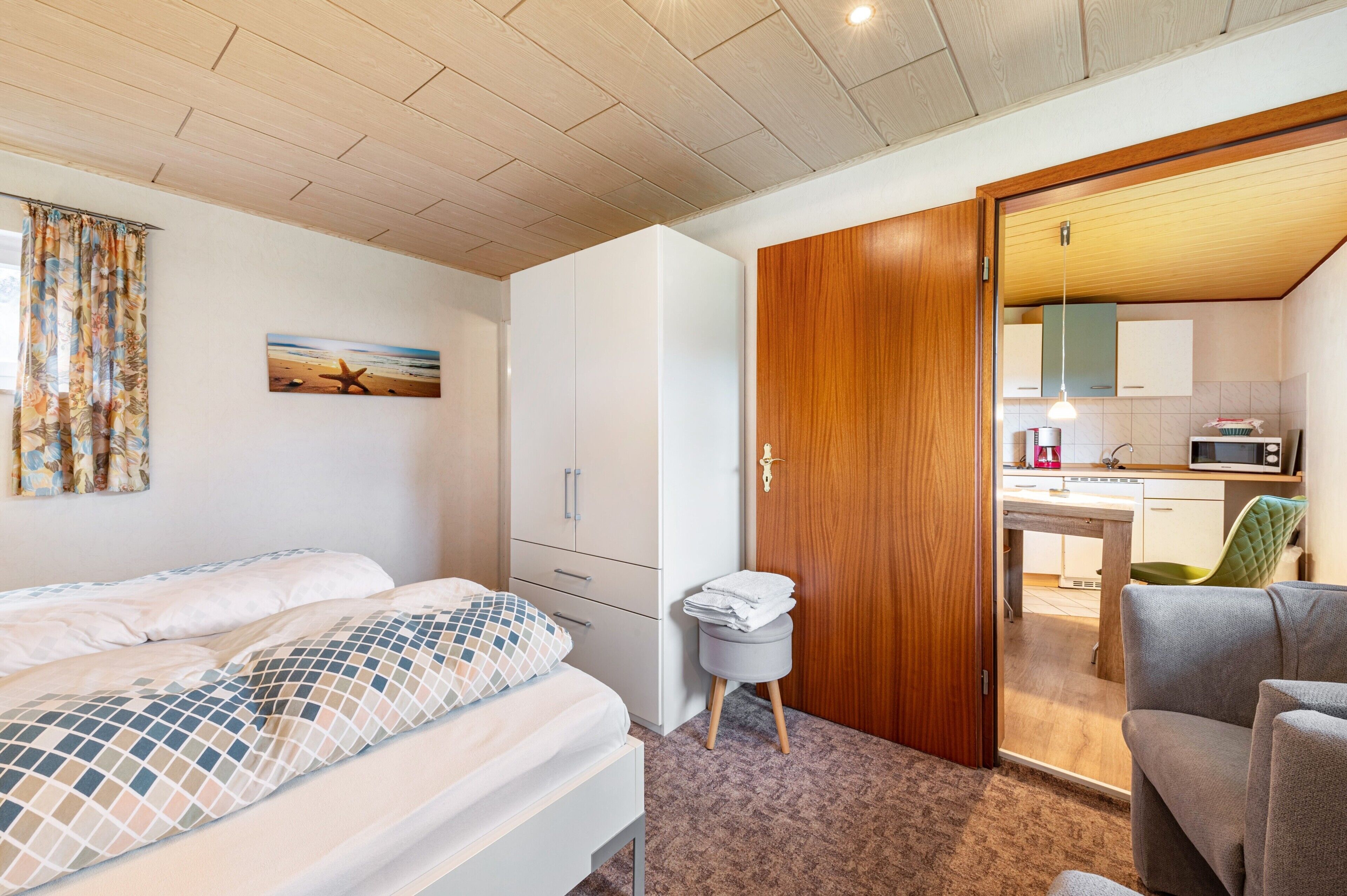 Un dormitorio con cama, armario, un taburete pequeño y vistas a una zona de cocina.
