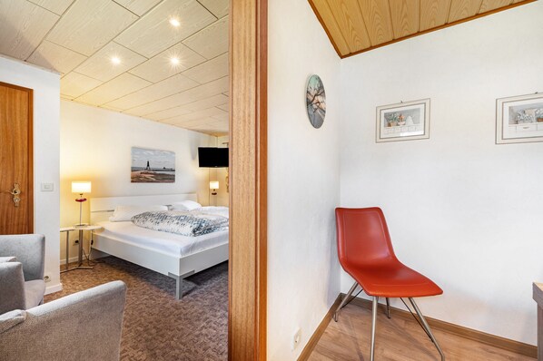 Habitación de hotel con cama, una silla roja, escritorio, lámpara y obras de arte en las paredes.